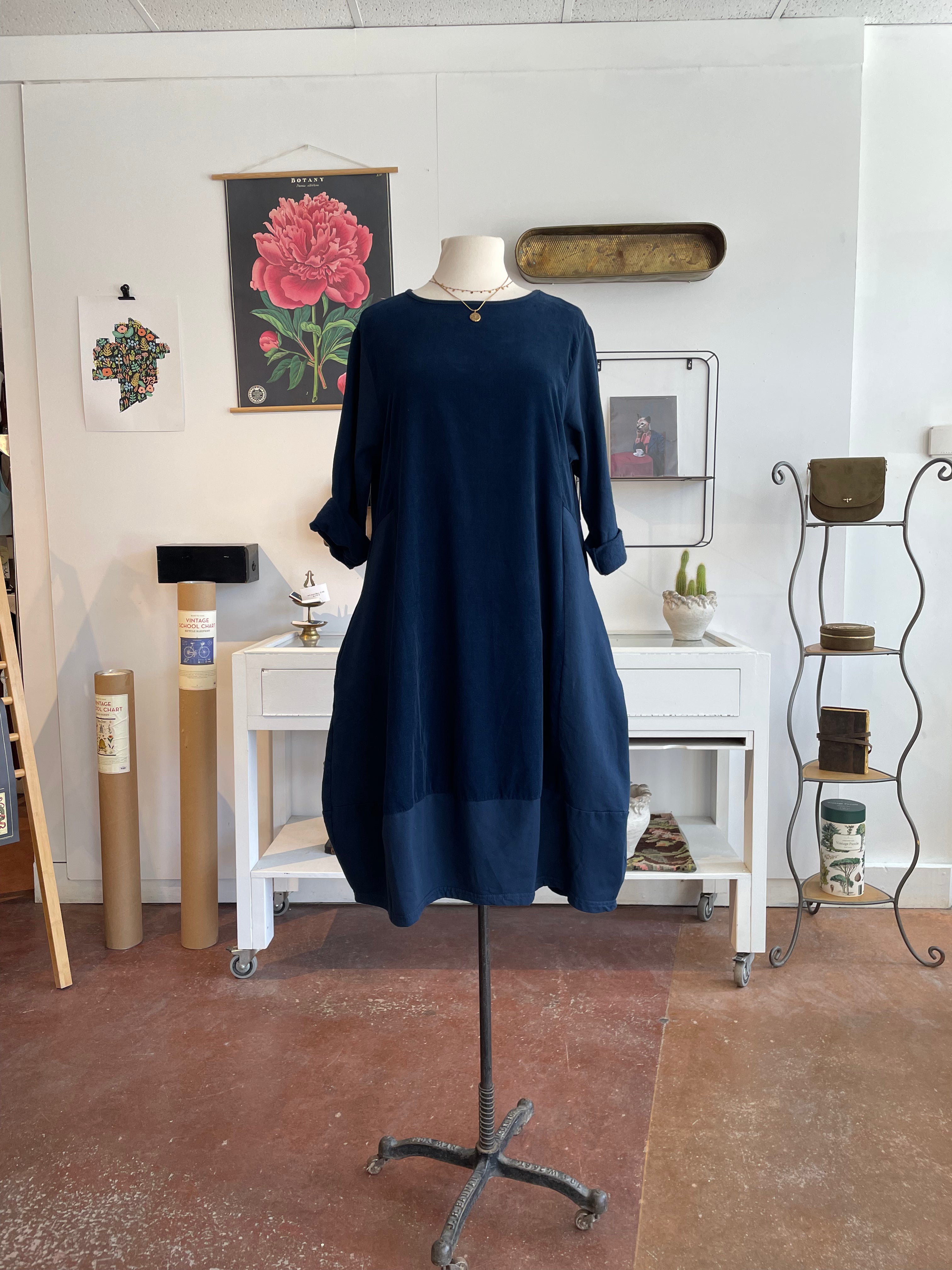 A navy blue long-sleeve tunic dress displayed on a mannequin torso stand in a styled studio or boutique interior with white walls, framed wall art, a small white desk, and shelving.
